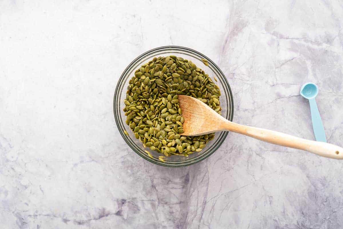 A small glass bowl filled with green seeds with a wooden mixing spoon with a blue measuring spoon
