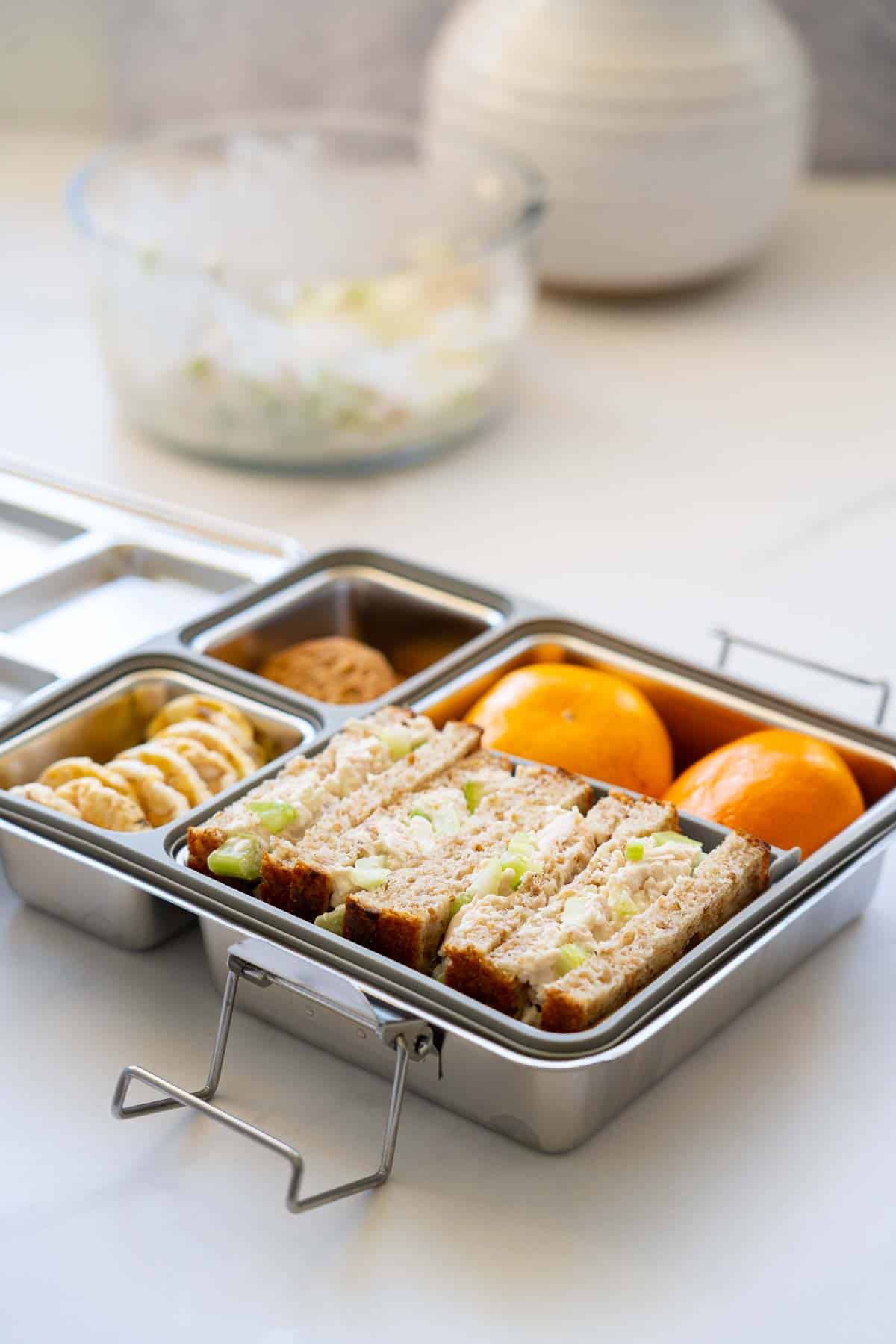 A stainless steel lunch box packed with fruit, crackers, biscuits and chicken salad sandwiches,
