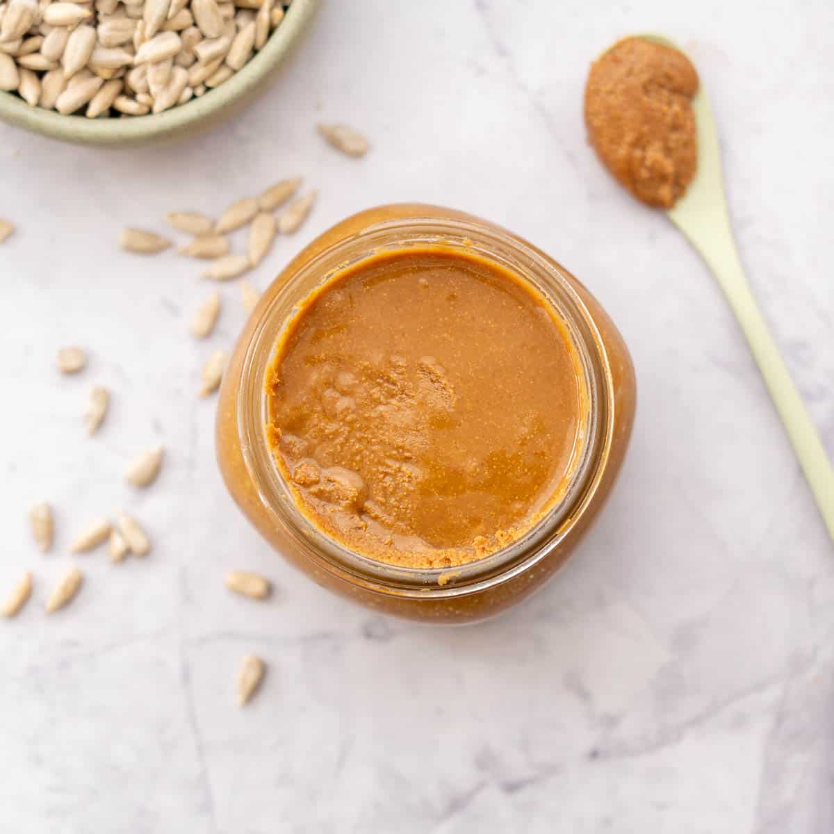 A jar of brown sunflower seed spread with a green spoon beside it and a bowl of raw sunflower seeds