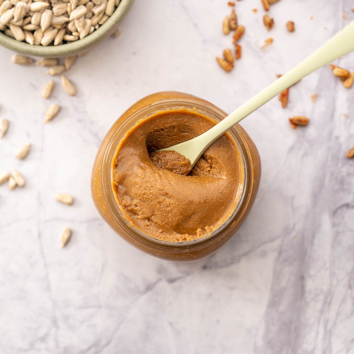 Jar of spread with a green spoon in the jar, with roasted and un-roasted seeds in the background.