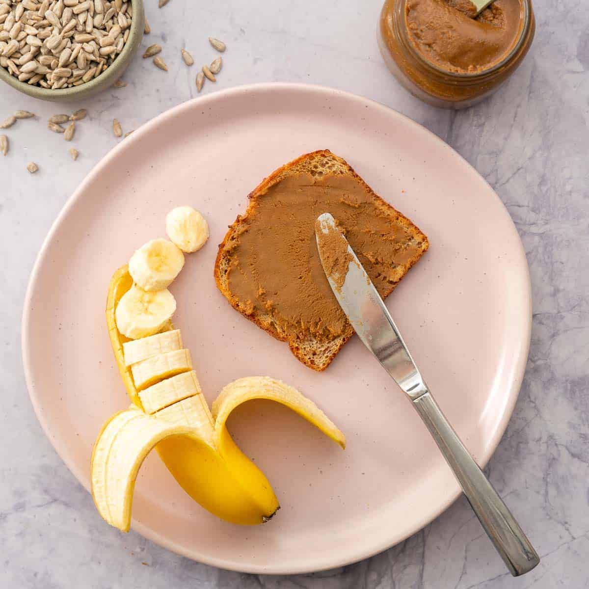 Pink plate with brown bread with sunflower spread next to a banana chopped up in the skin.