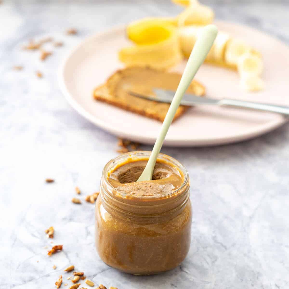 In front a small jar with sunflower seed butter with a green spoon in the jar. Faded in the background is a pink plate with a piece of bread with spread on it with a knife and chopped banana beside the bread