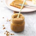 A glass jar of seed butter on a marble bench top with a slice of toast and sliced banana in the background.