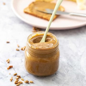 A glass jar of seed butter on a marble bench top with a slice of toast and sliced banana in the background.
