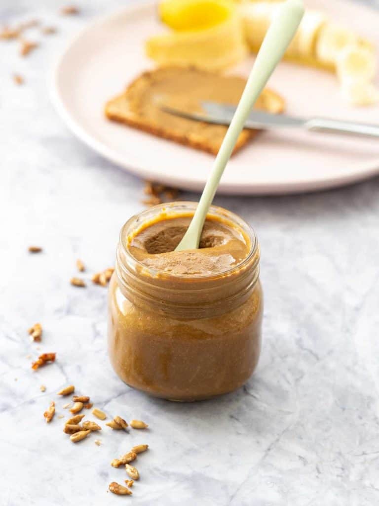 A glass jar of seed butter on a marble bench top with a slice of toast and sliced banana in the background.