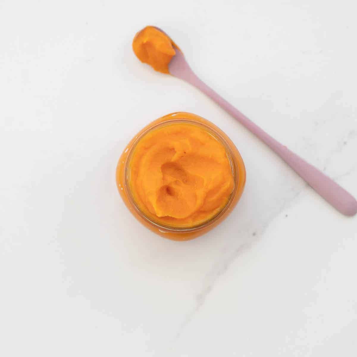 Looking down into a glass jar of orange vegetable puree on a bench top with a spoonful of puree lying next to the jar.