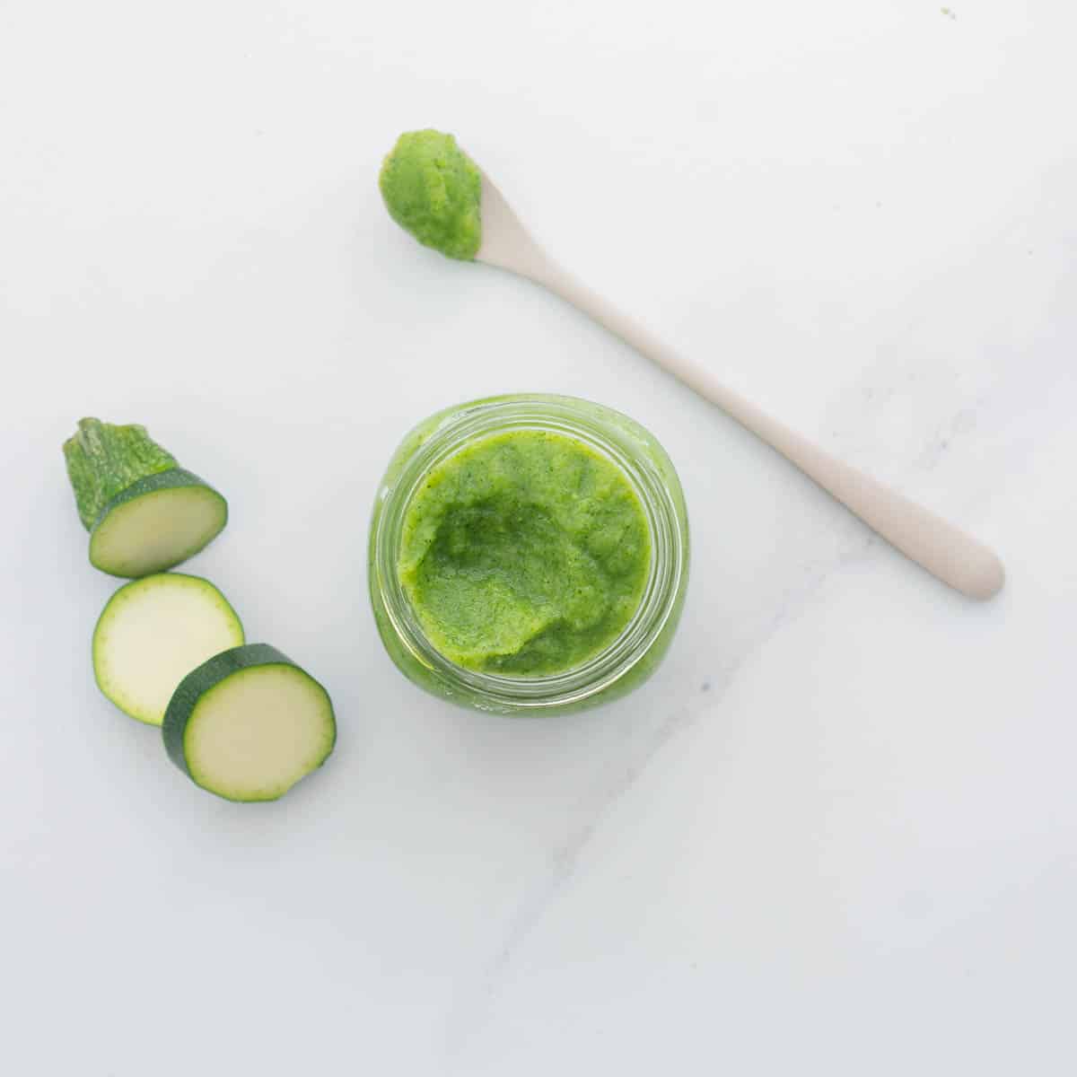 A jar of green vegetable puree on a bench next to slices of zucchini and a bamboo spoon.