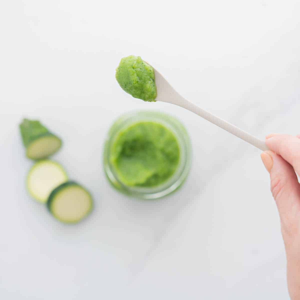 A spoonful of zucchini puree being held above a jar of puree and three zucchini slices on a marble bench 