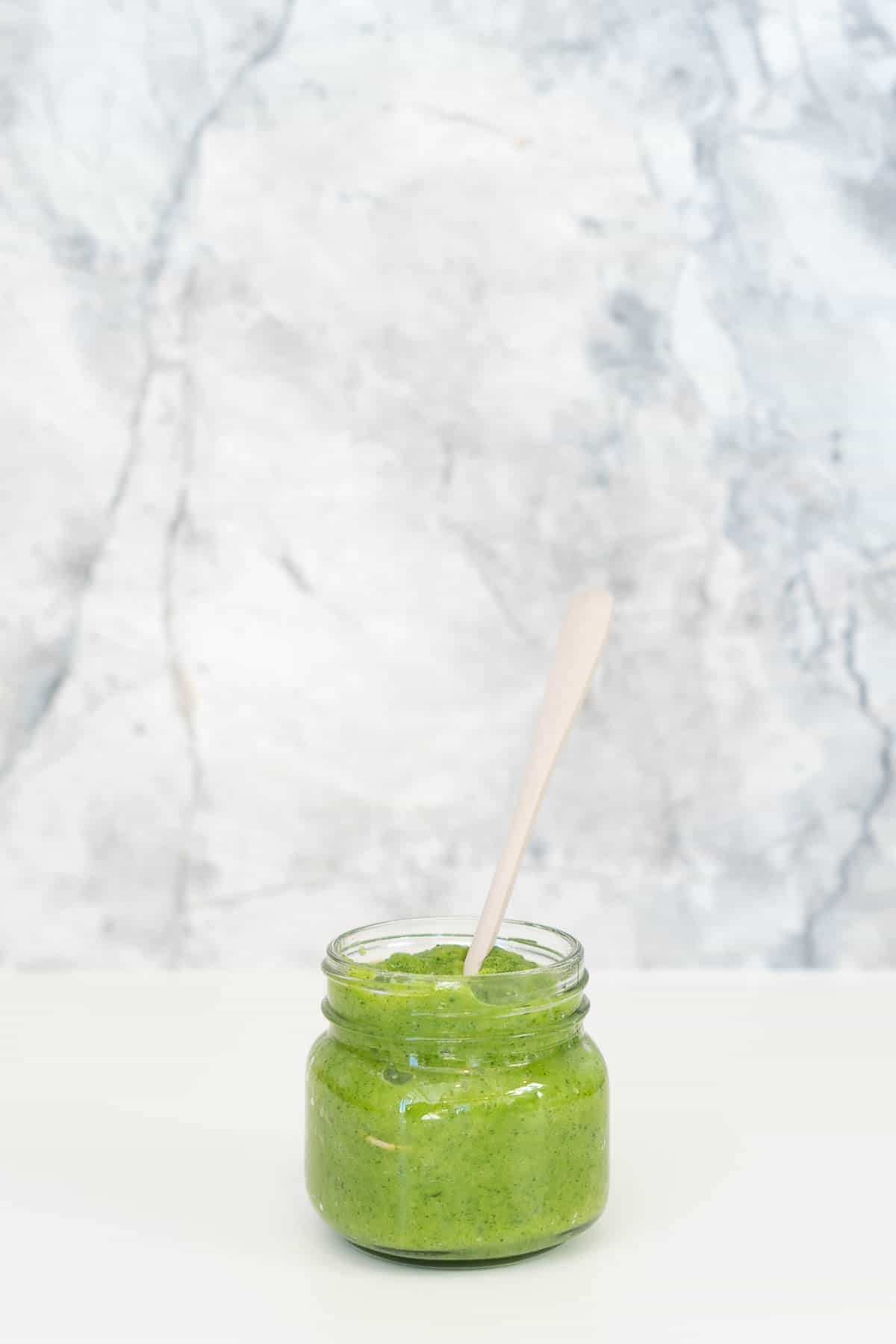 A glass jar of green zucchini puree sitting in front of a grey marble splashback.