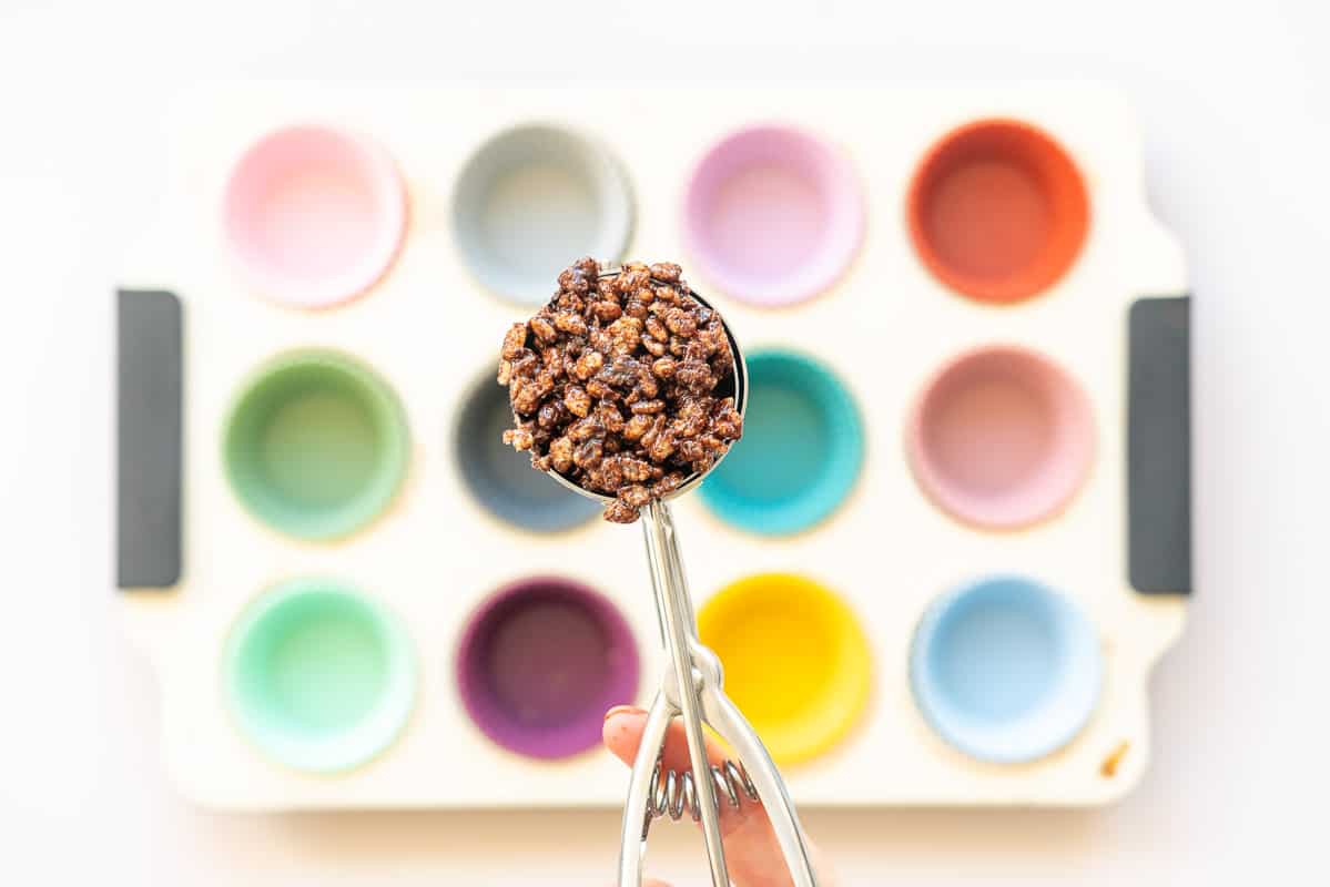 An ice cream scoop of chocolate crackle mix being held above a muffin tray.