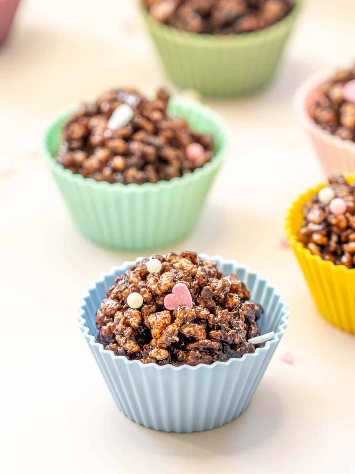 Chocolate crackles in multi coloured reusable silicone cupcake liners on a white bench top scattered with heart shaped sprinkles.