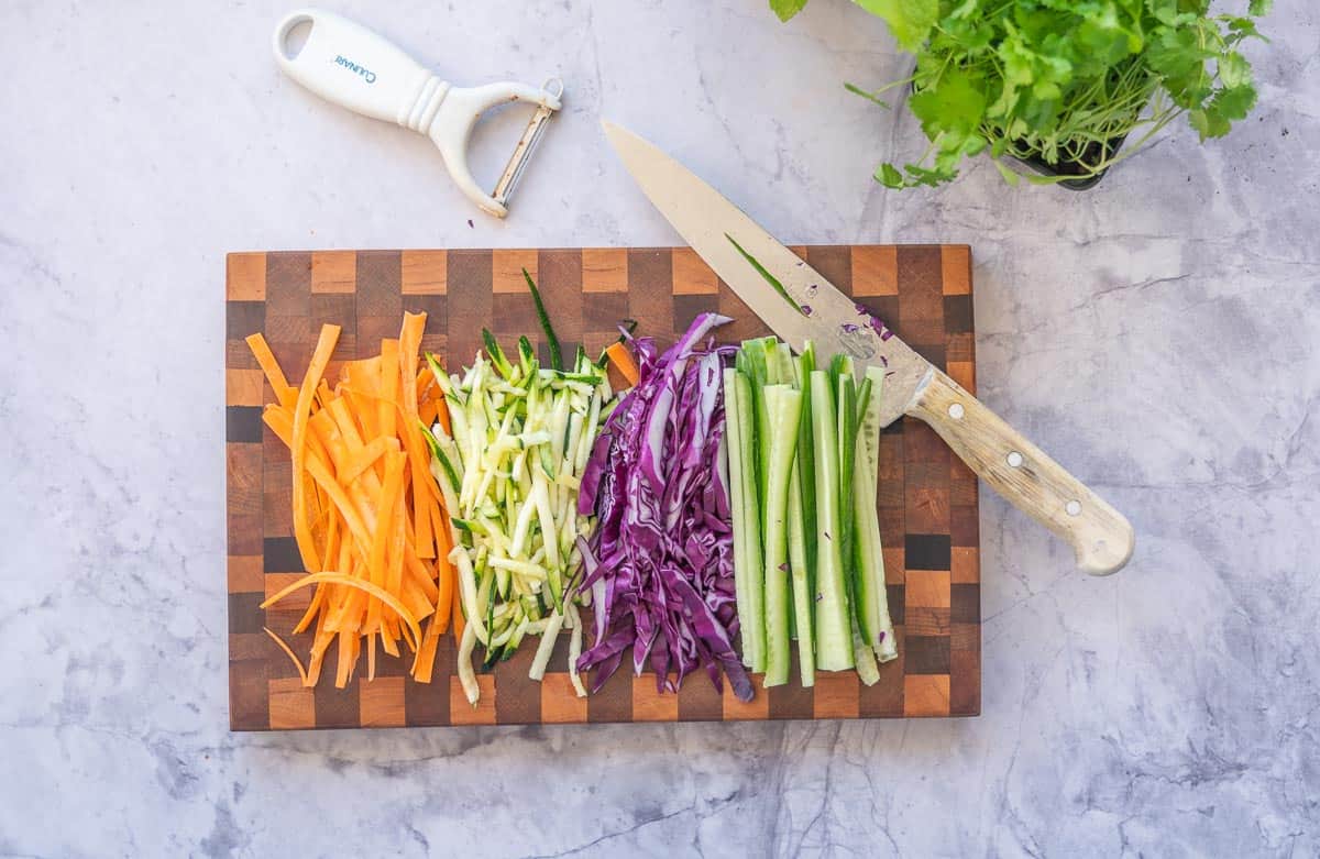 Sliced carrot, zucchini, purple cabbage and cucumber on chopping board with peeler and knife.