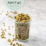 A jar of roasted pepitas on a marble bench, some pepitas are scattered on the bench top with text overlay.