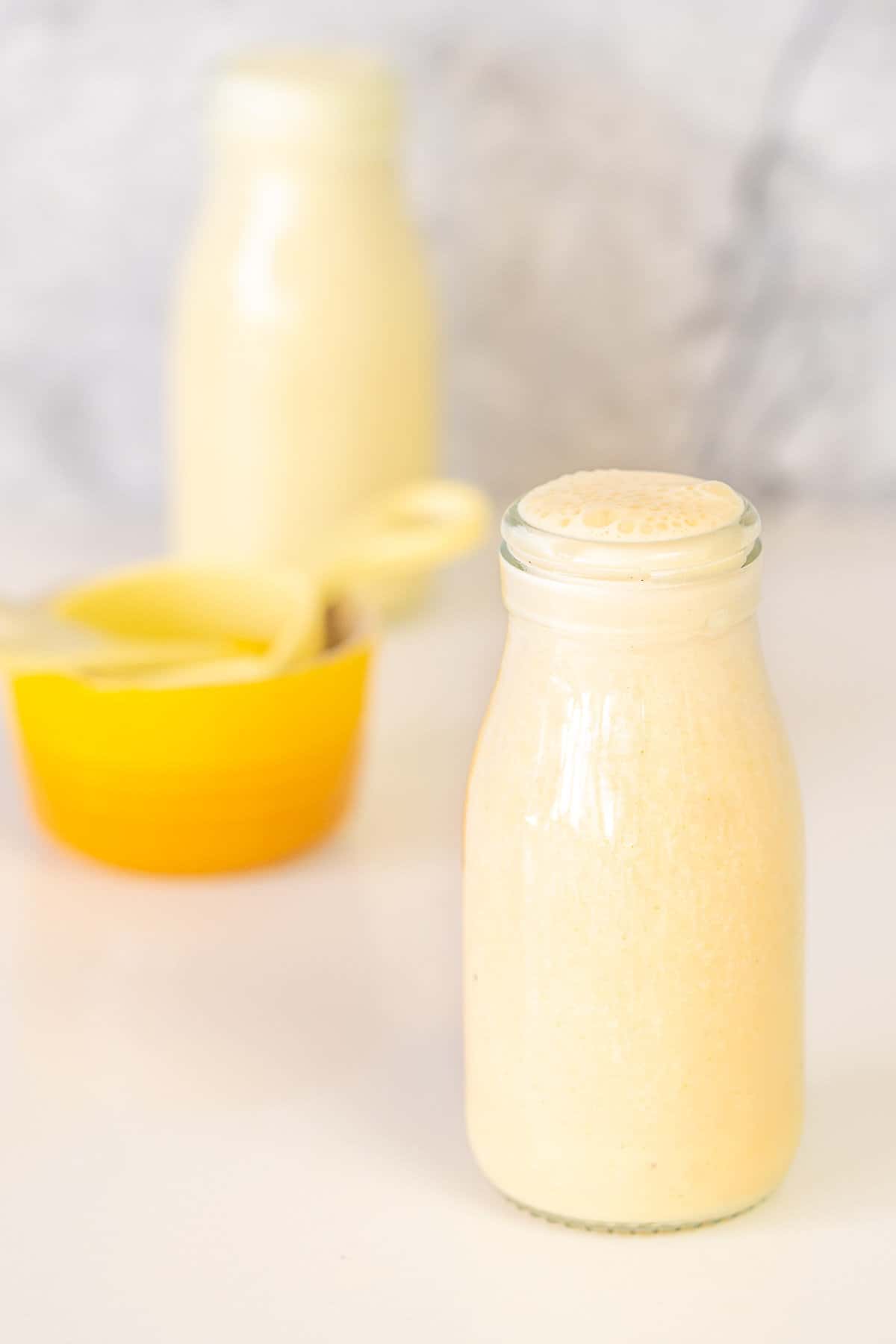 A small glass milk bottle filled with banana milk next to a yellow ramekin and yellow measuring cup.