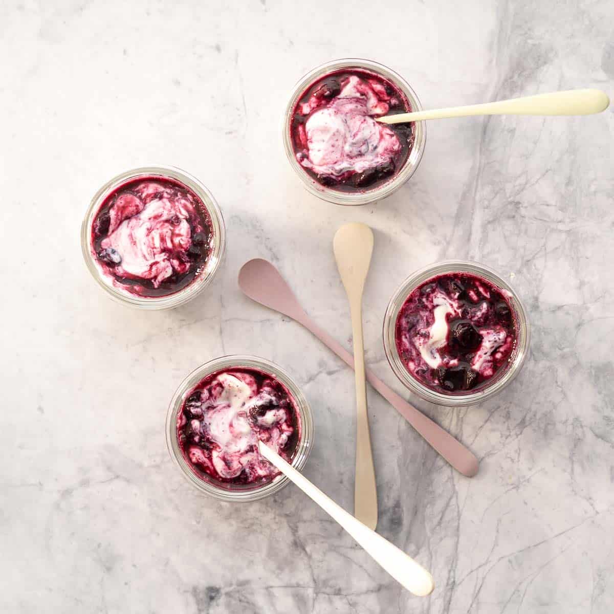 Four bowls of blueberry yogurt on a marble bench top with colourful spoons.