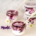 Blueberry yogurt in small glass jars on a bench top scattered with white and purple flowers.