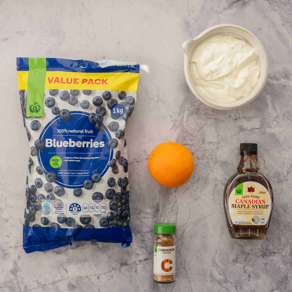 Frozen blueberries, orange, cinnamon, maple syrup and yogurt laid out on a bench top.