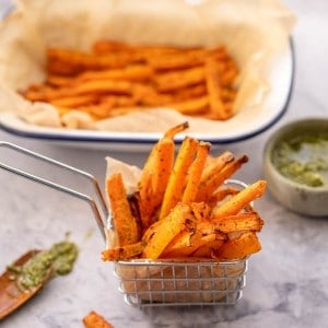 A small metal basket filled with carrot fries on a bench next to a spoonful of green pesto.