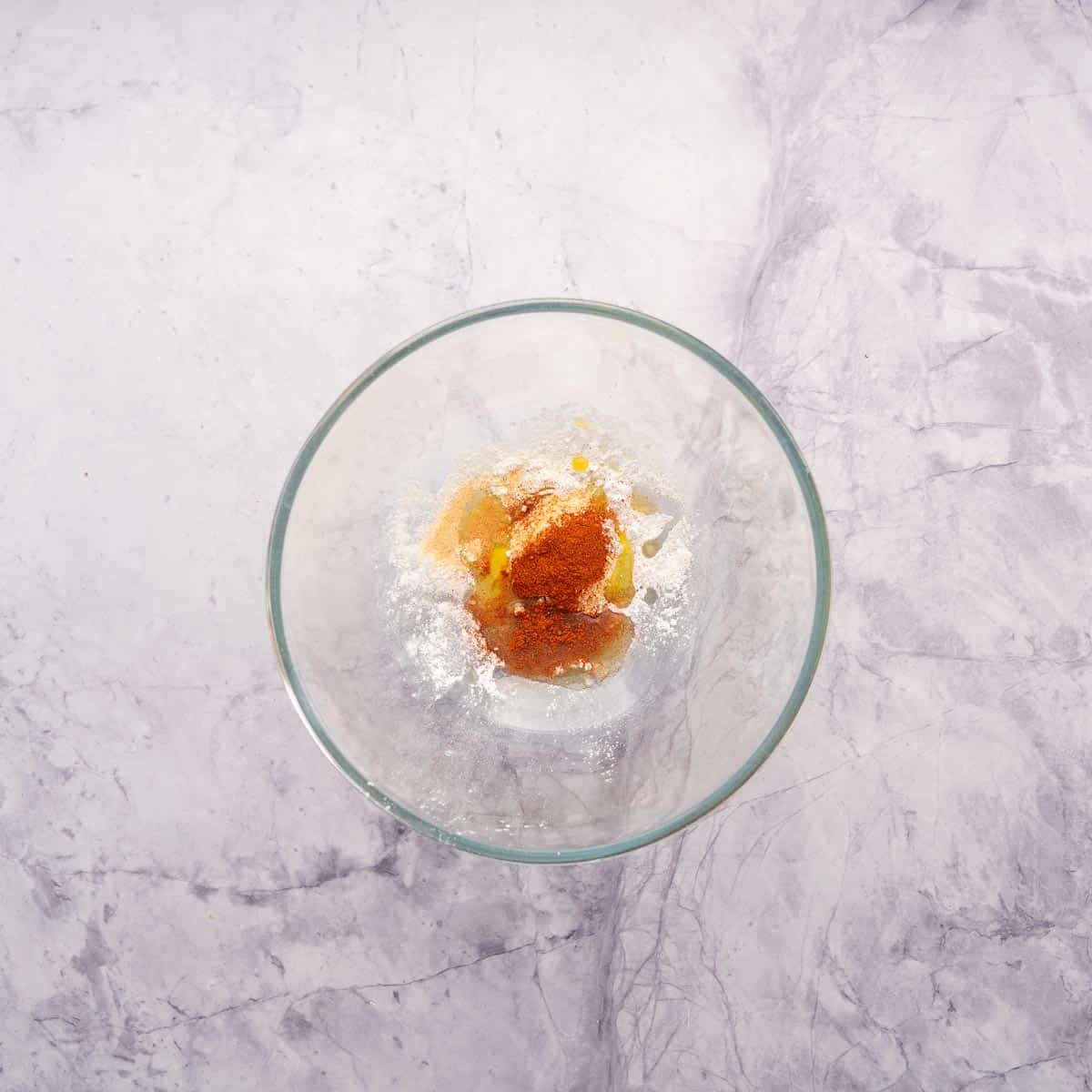 A large glass mixing bowl with oil, corn flour and spices in the bottom of it.