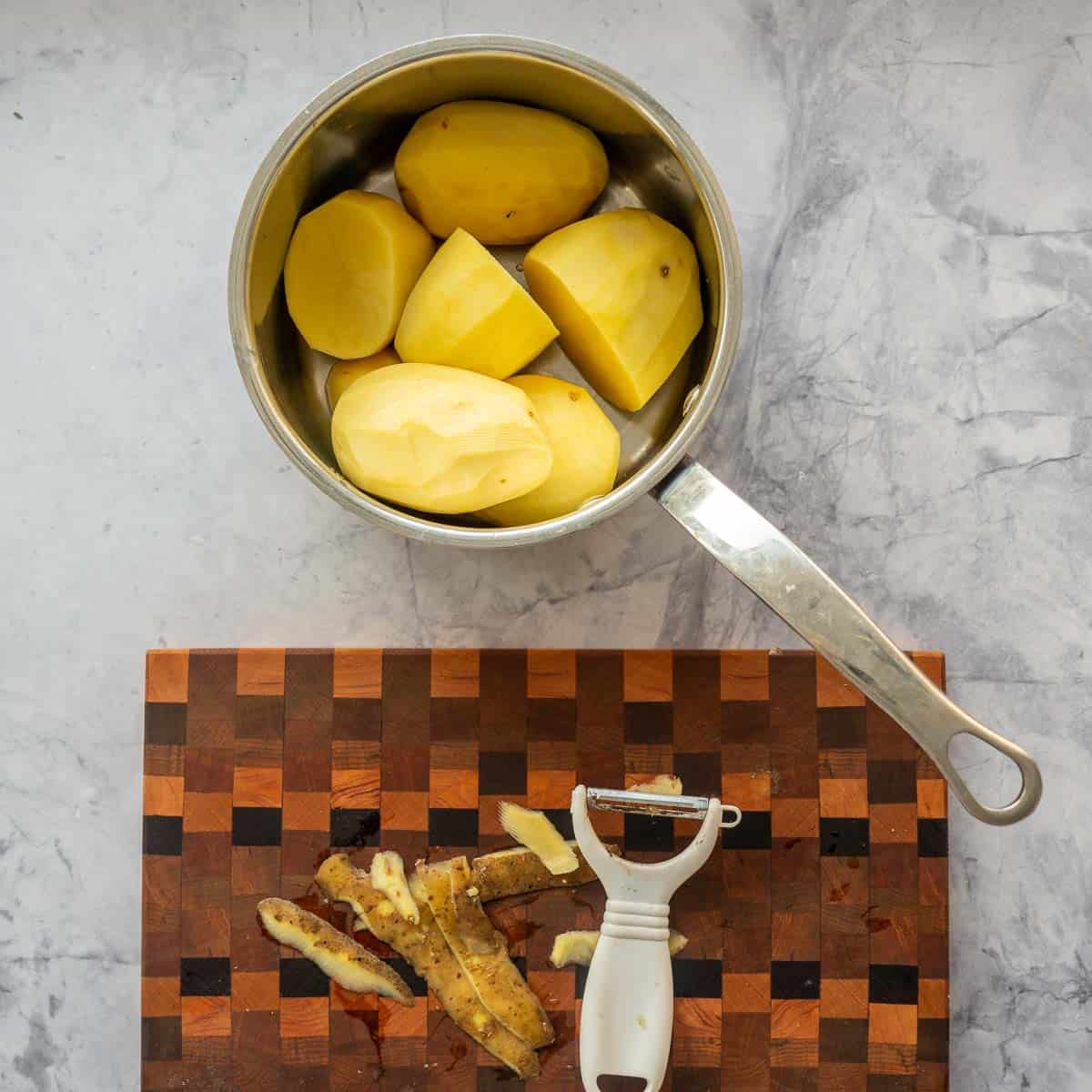 Peeled potatoes in a pot next to a chopping board with a peeler and potato peel.