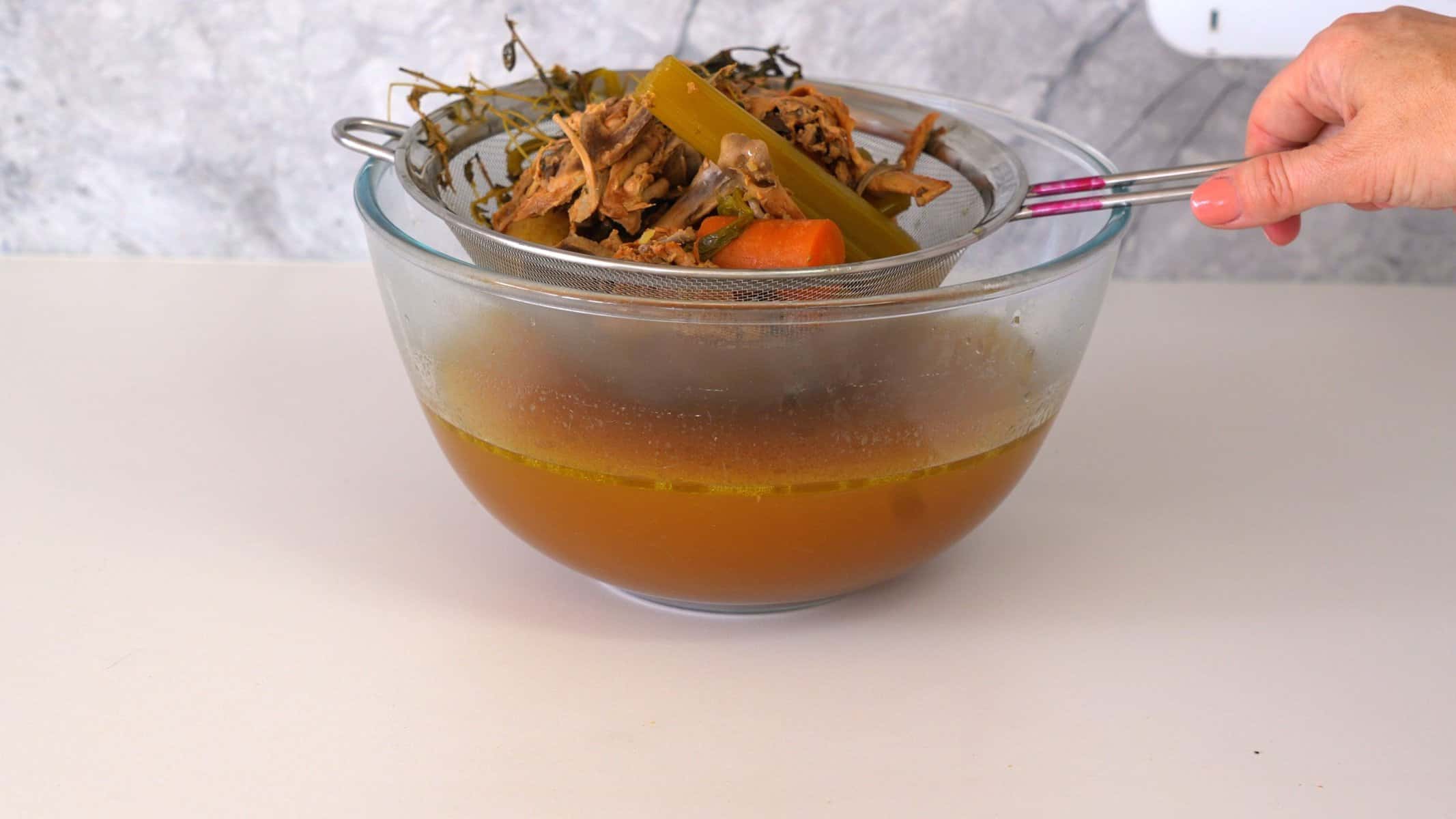 A large bowl sitting on the bench with drained bone broth inside of it, with a hand holding a sieve full of cooked vegetables and bones.