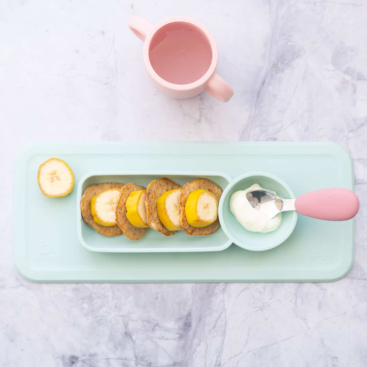 Banana slices, pancakes and yogurt on a mint green baby plate with a pink handled spoon and pink sippy cup.
