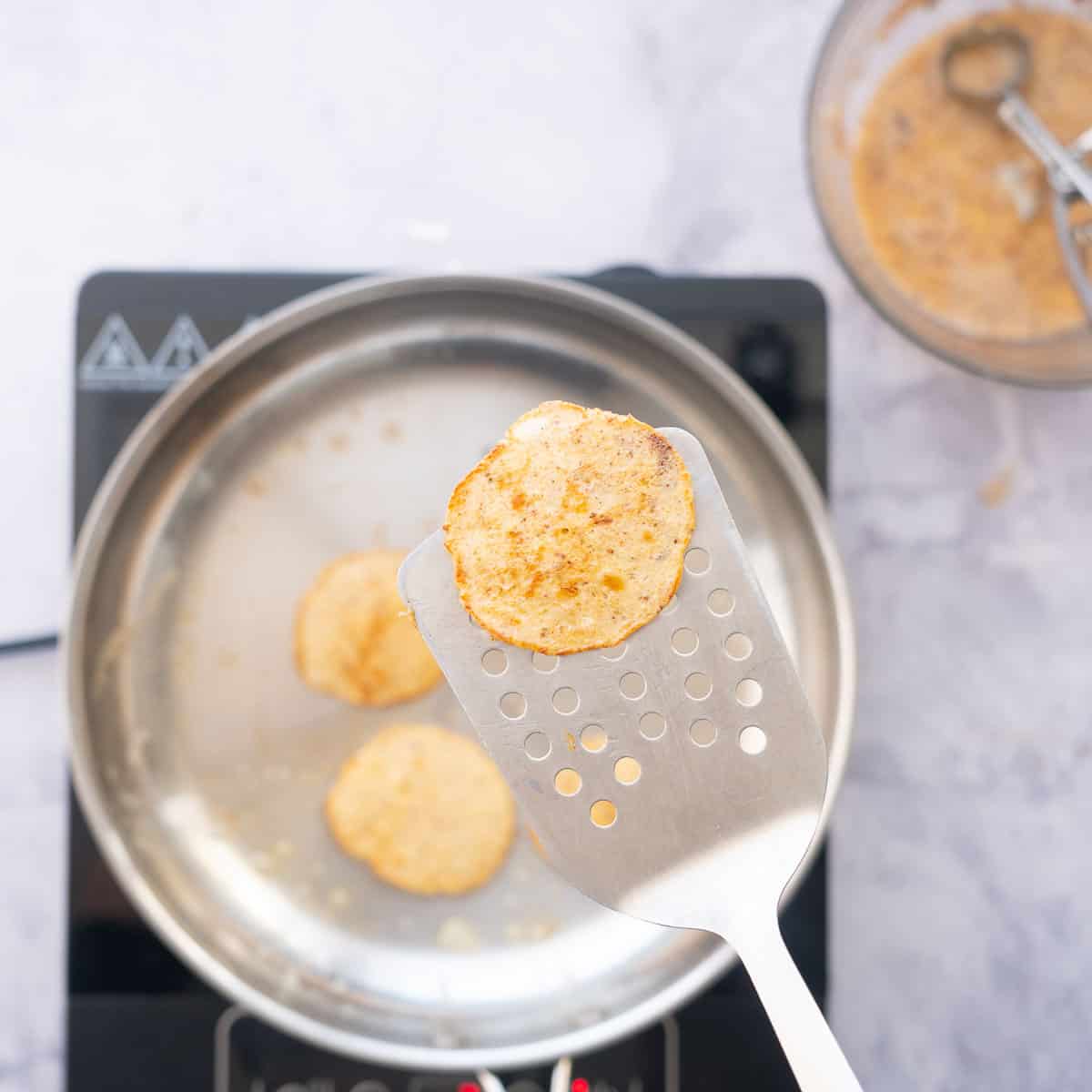 A small golden cooked pancake being lifted on a stainless steel spatula.