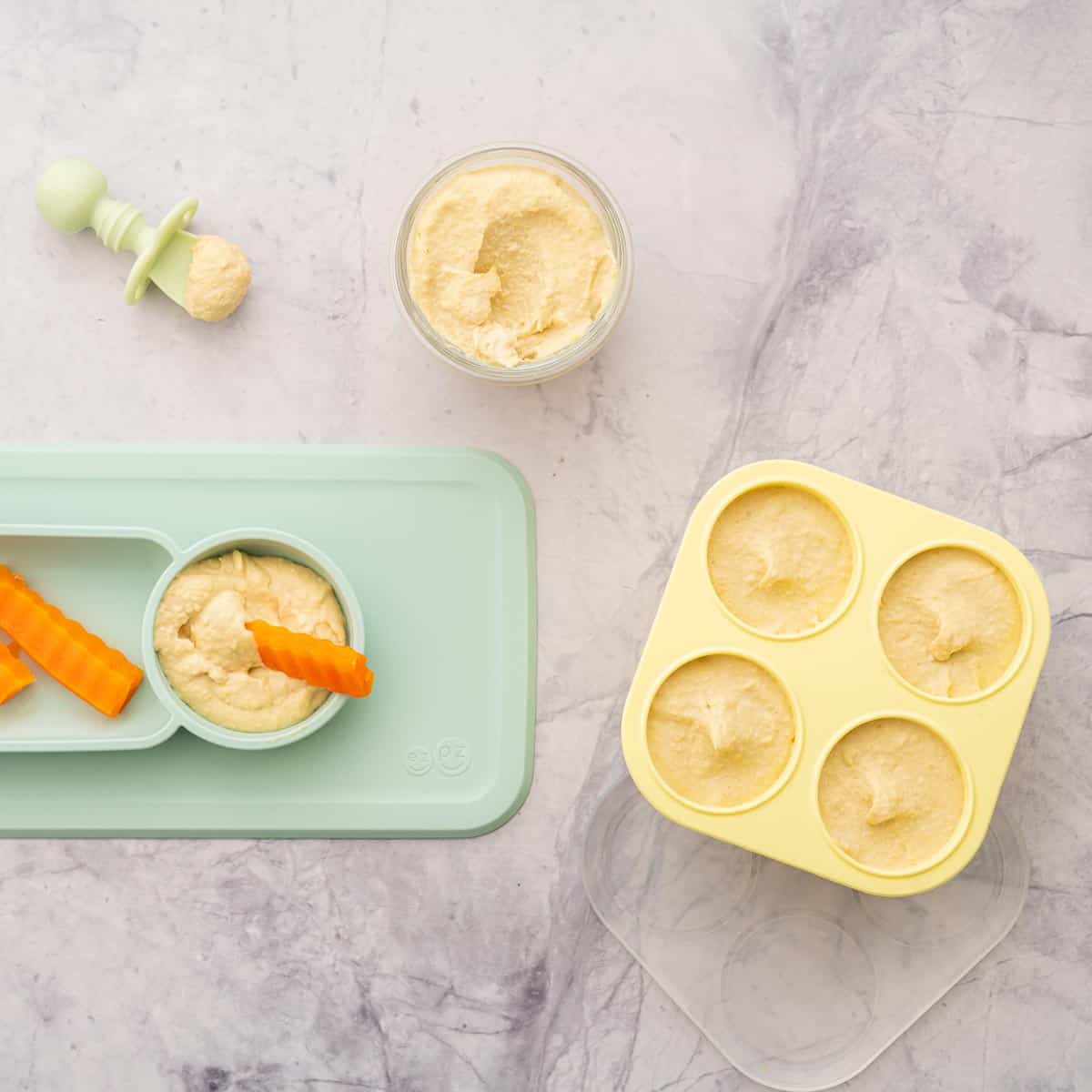 Yellow muffin cases filled with baby hummus sitting next to a ramekin of hummus and a baby plate with carrots dunked into hummus and a baby spoon with hummus on it.