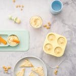 Yellow muffin cases filled with baby hummus sitting next to a ramekin of hummus and a baby plate with carrots dunked into hummus, chickpeas scattered on the bench next to a baby spoon and cup of water and a plate with toast with hummus spread