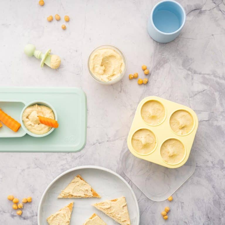 Yellow muffin cases filled with baby hummus sitting next to a ramekin of hummus and a baby plate with carrots dunked into hummus, chickpeas scattered on the bench next to a baby spoon and cup of water and a plate with toast with hummus spread