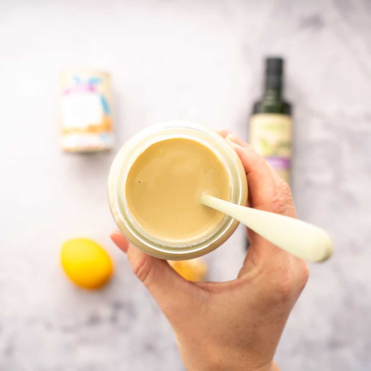 a hand holding up a jar of tahini with a spoon inside of it, hovering above the bench with the laid out ingredients below it.