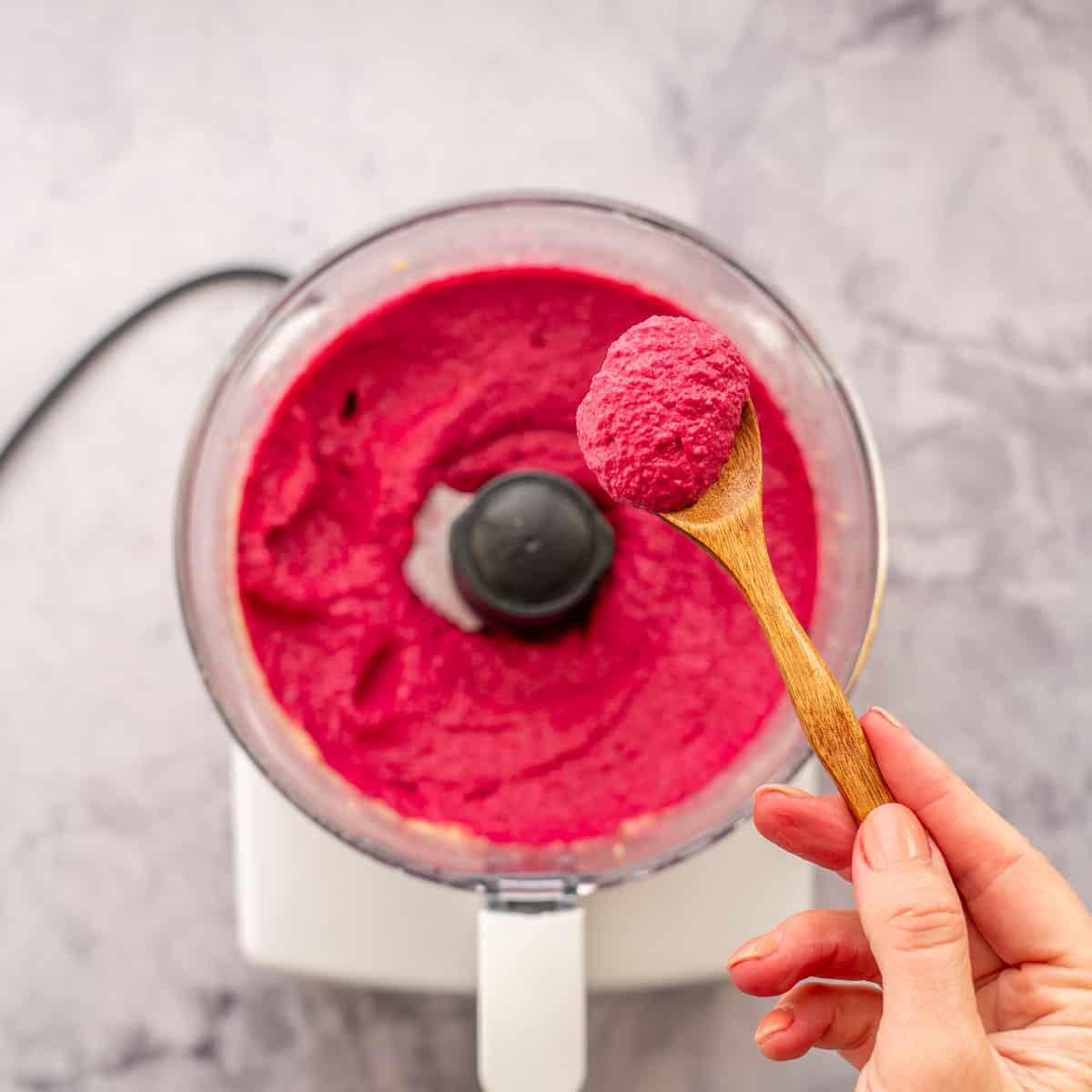 A food processor full of whipped beetroot hummus with a hand holding a spoonful above it.