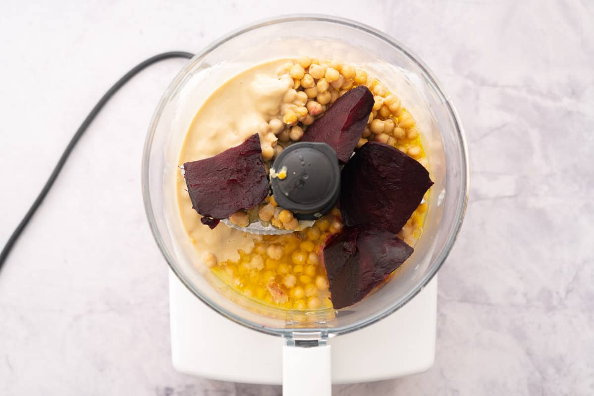 A food processor filled with chickpeas, roasted beetroot, tahini and olive oil sitting on a bench.