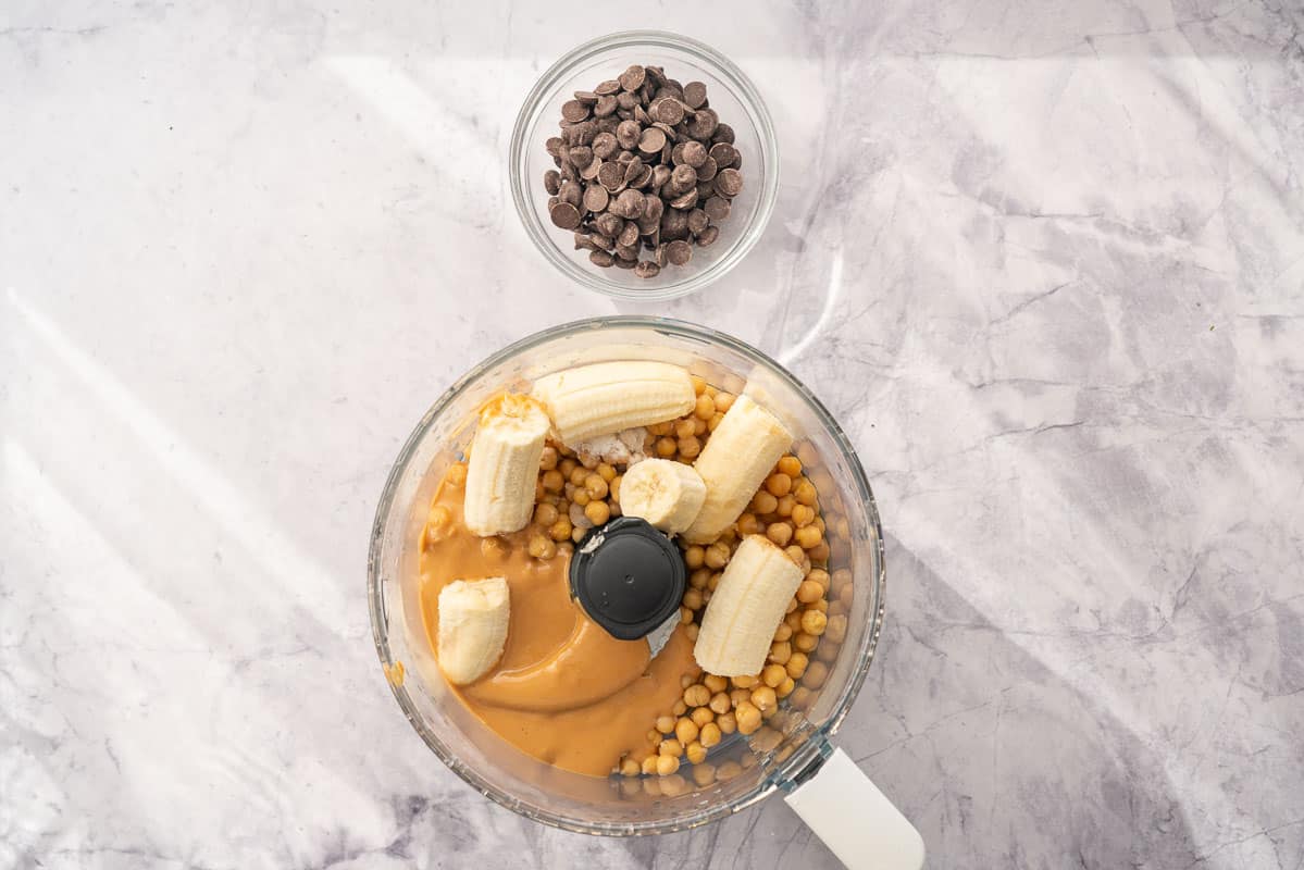 a food processor sitting on the bench with bananas, peanut butter, chickpeas inside of it, sitting next to a ramekin on chocolate chips
