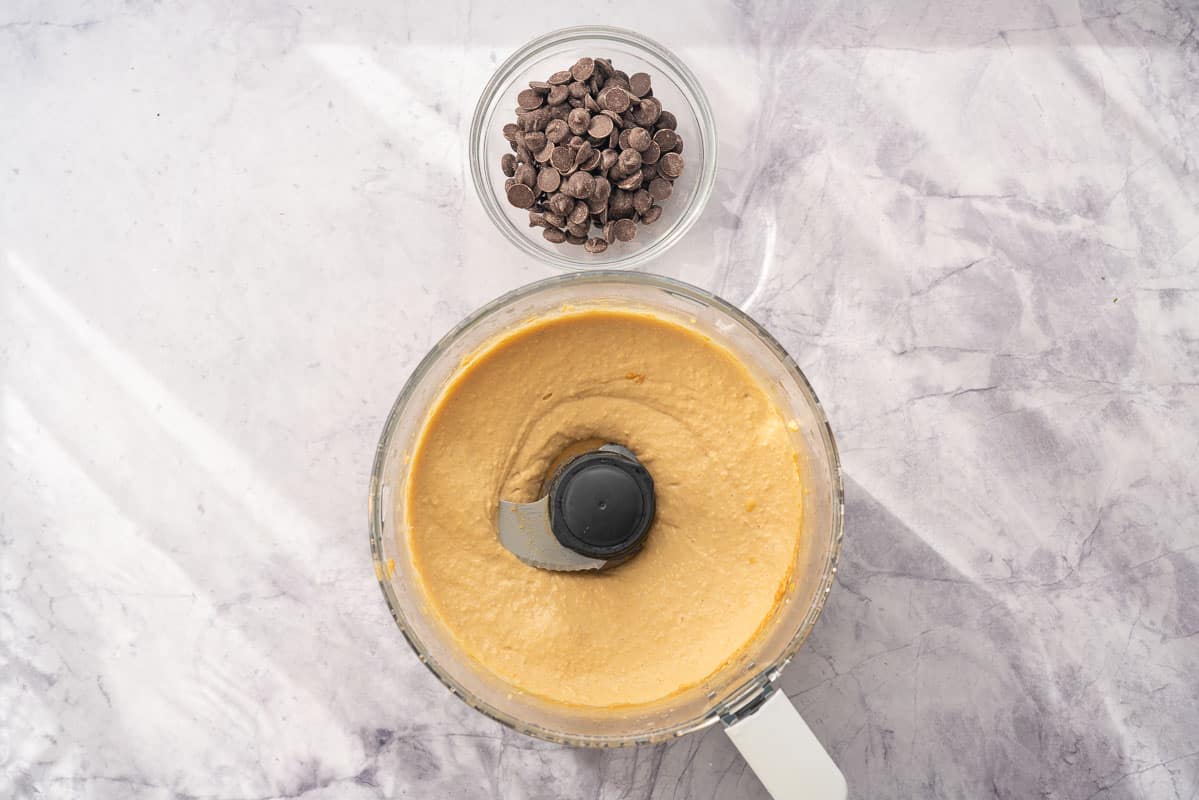 A food processor sitting on the bench with ingredients whizzed together inside of it, sitting next to a ramekin of chocolate chips