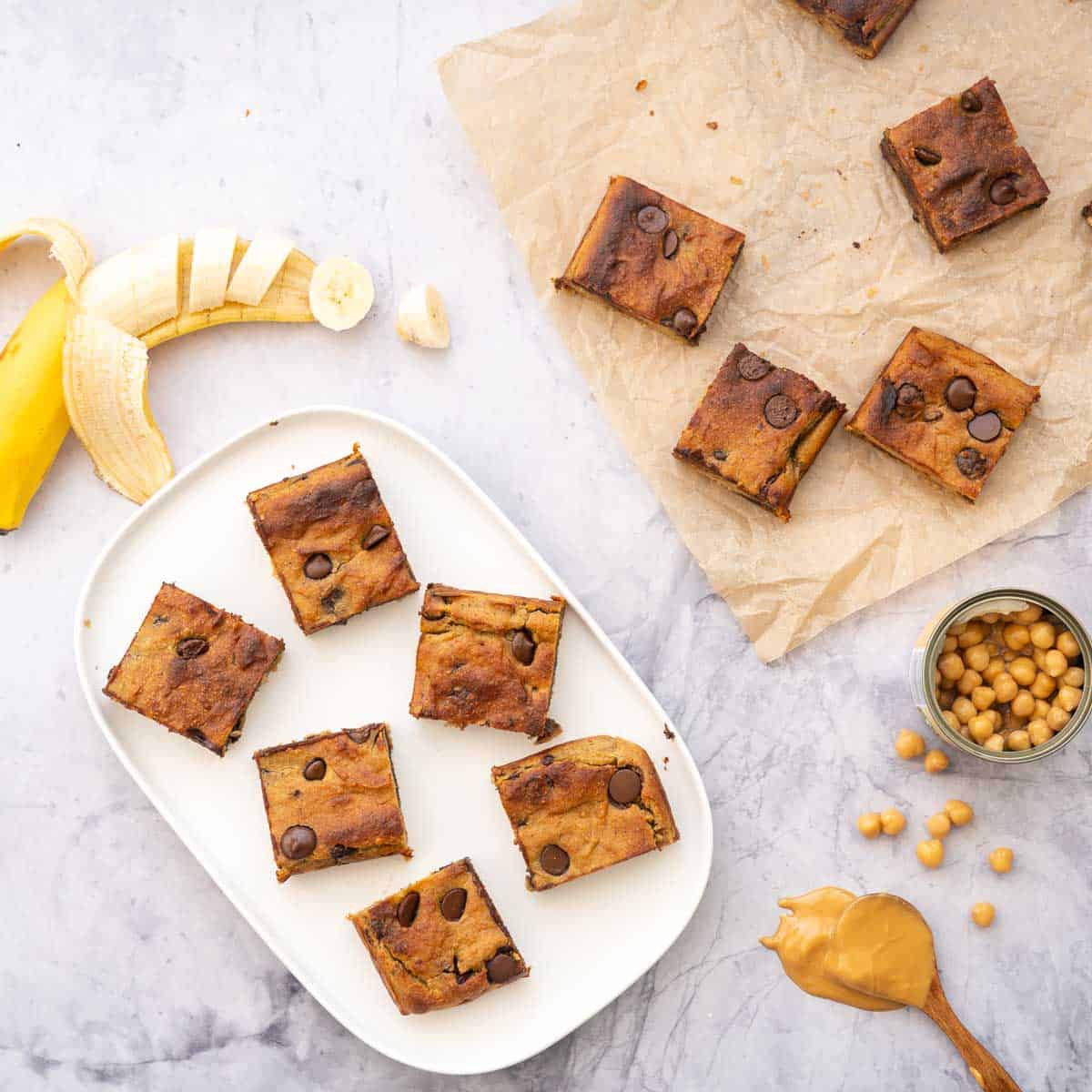 Baked Chickpea Blondies sliced and sitting on brown crinkled baking paper and some sitting on a white plate next to a sliced banana and a small can of chickpeas and peanut butter drizzling of a spoon