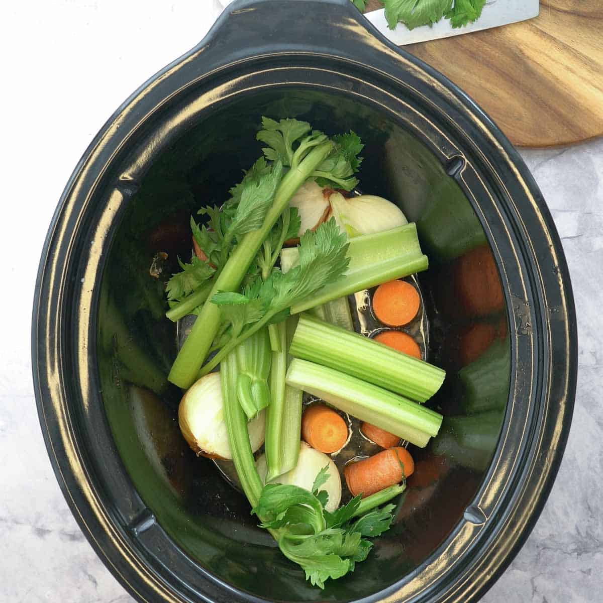 A large croc pot sitting on a bench, with chopped up carrots, celery and onions inside of it.