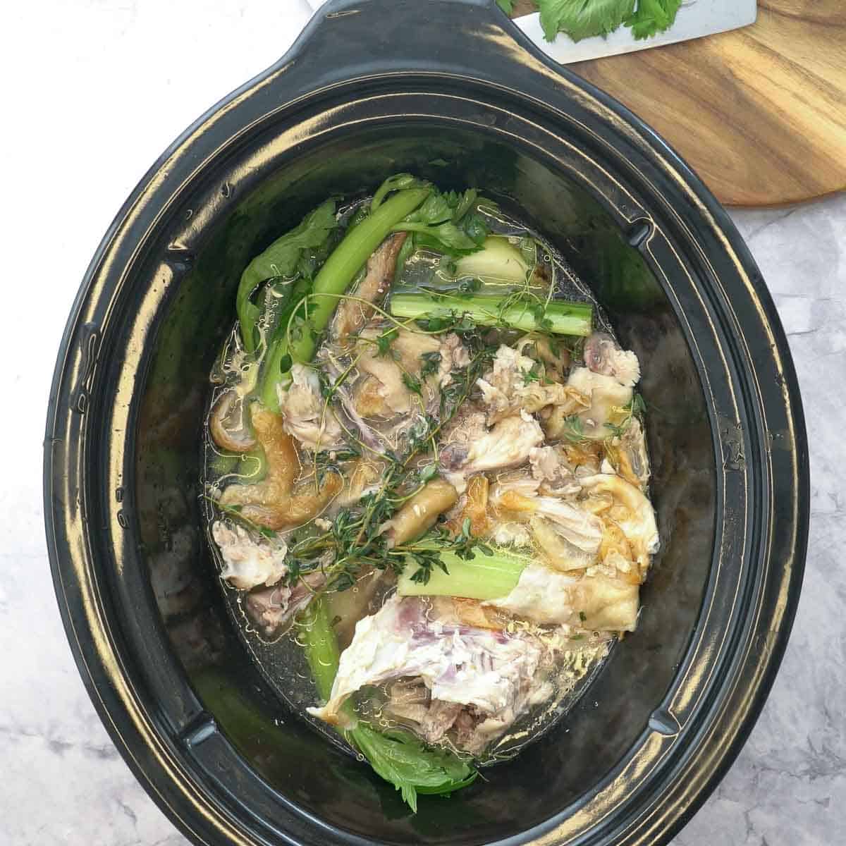 A croc pot sitting on the bench with chopped up vegetables, chicken bones and sprigs of thyme covered in water.