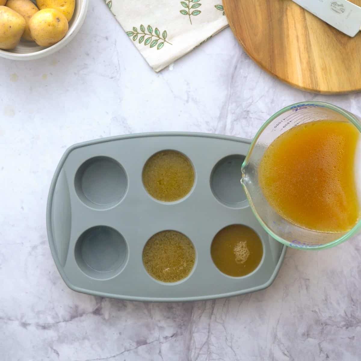 A jug full of bone broth being poured into a 6 case muffin tin sitting on a bench with a chopping board and a tea towel next to it.
