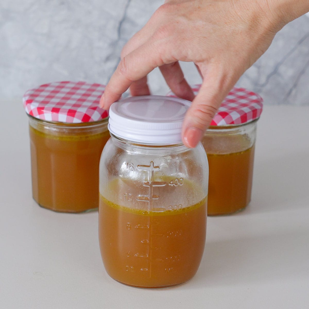 3 mason jars sitting full of bone broth sitting the bench with a hand screwing the lid onto one of them.