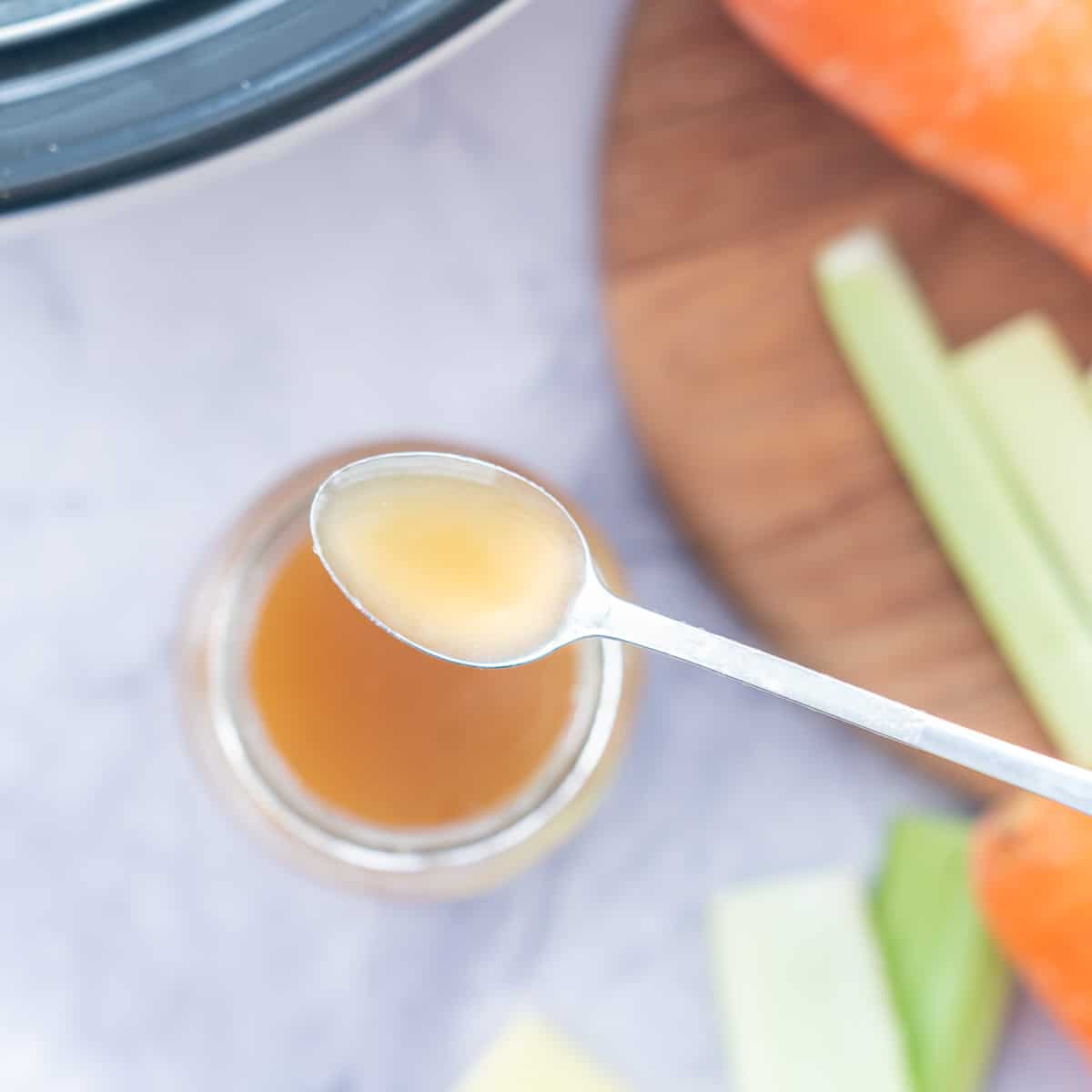 A spoon full of bone broth being held above a a full jar full of broth with a chopping board and vegetables laid out next to it.