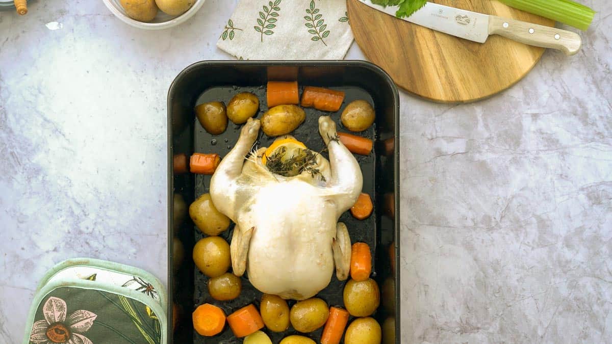 A large black roasting dish on a bench filled with roasted potatoes and carrots and a whole lemon and thyme stuffed chicken sitting amongst them. Next to it, is a chopping board with a large knife and celery stick, a oven mit and a small bowl of potatoes.