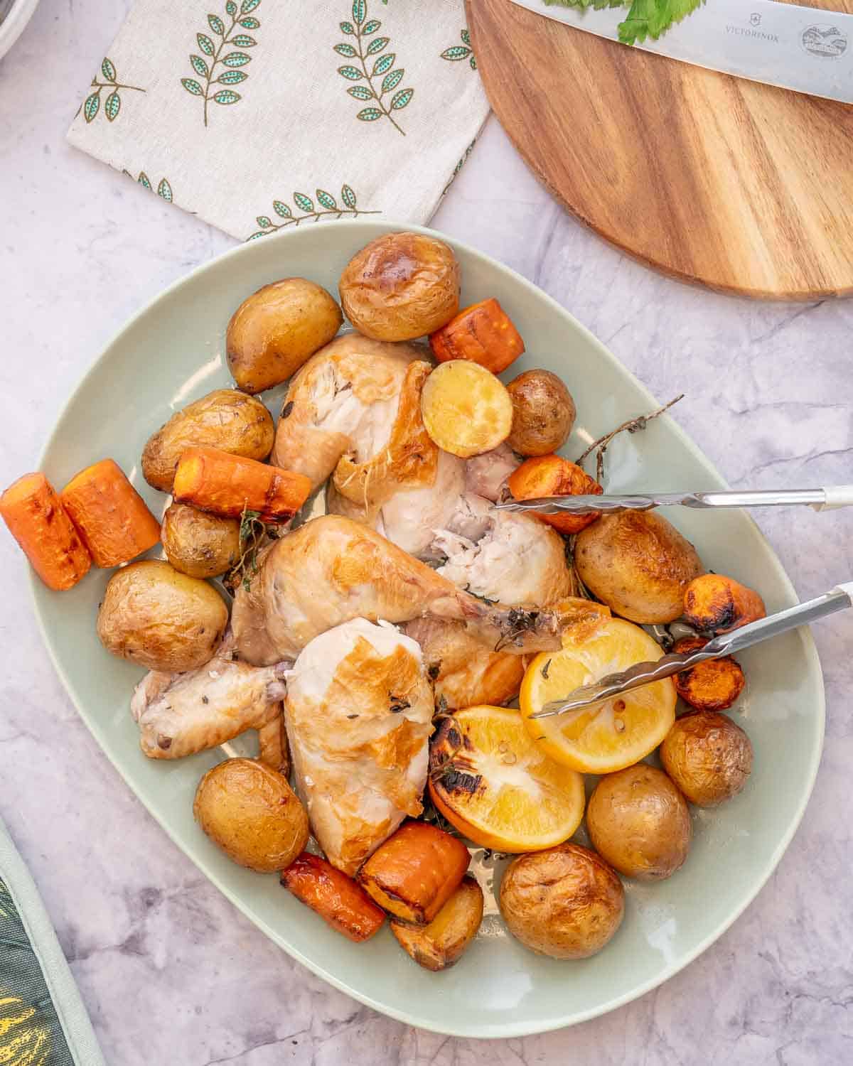 A large serving dish with tongs filled with roasted potatoes, carrots and chicken sitting on the bench next to a chopping board and knife and a tea bowl.