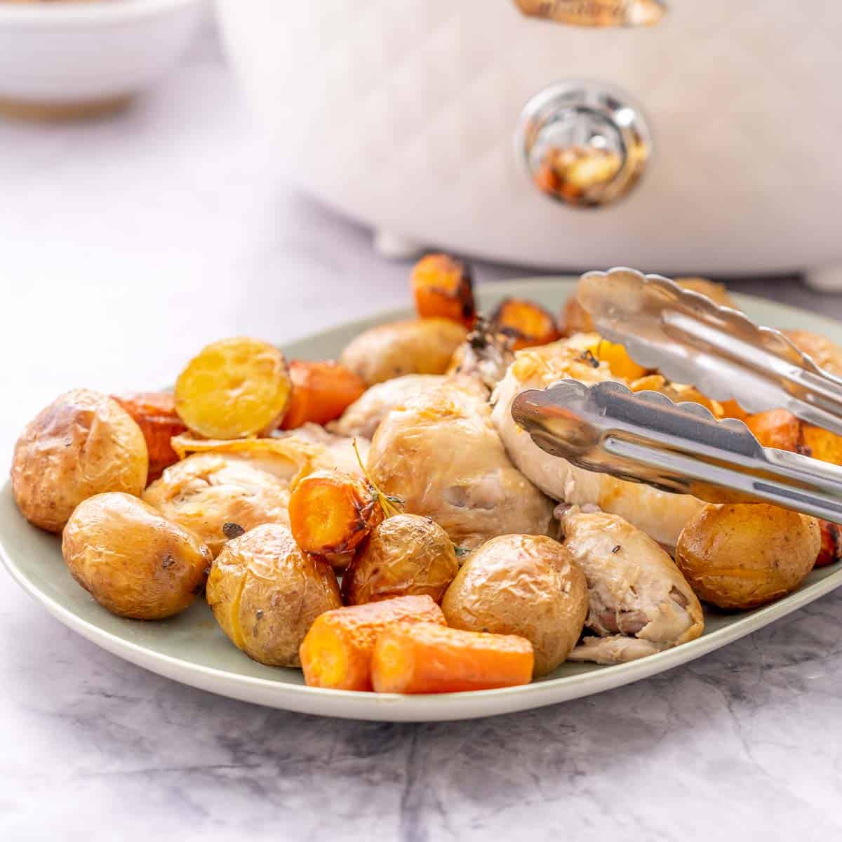 A large serving dish filled with roasted potatoes, carrots and chicken sitting on the bench in front of the croc pot with tongs about to dig in.
