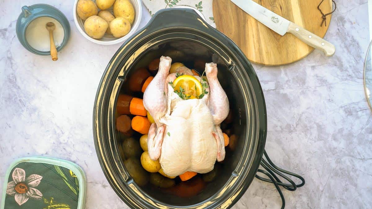A large crockpot sitting on the bench, with baby potatoes, chopped carrots and whole chicken inside of it, sitting next to a jar of salt, a cutting board with a knife on it and a small bowl of potatoes.