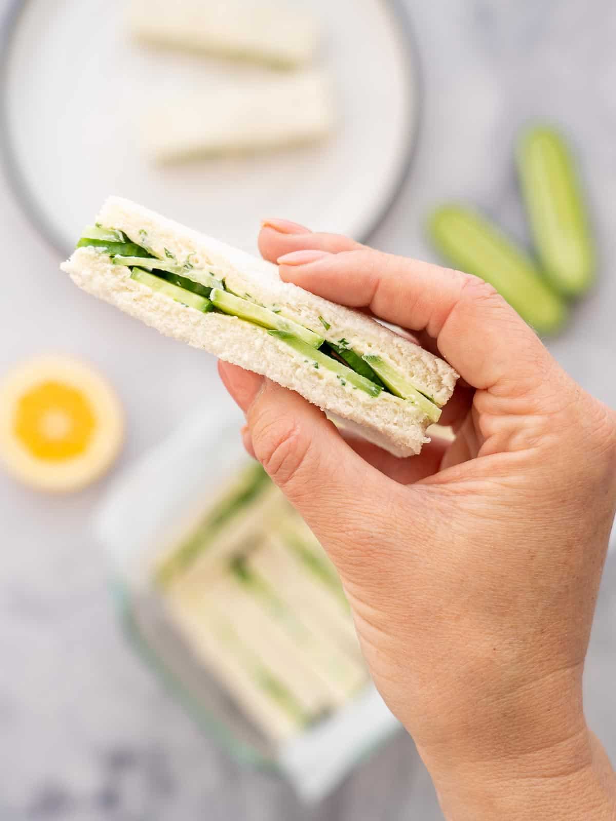 Hand holding a cucumber sandwich with sandwiches blurred in background.