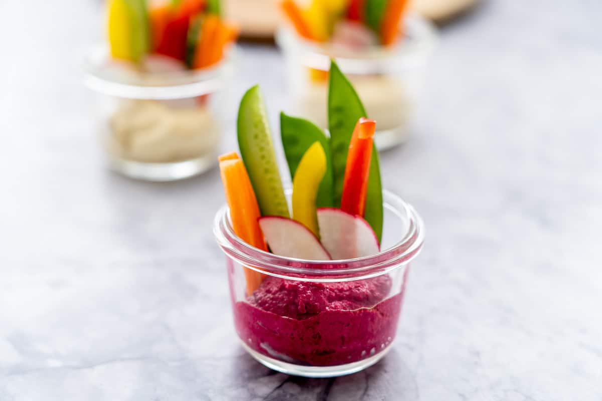 Three glass ramekins half filled with hummus and sliced pieces of radishes, carrots, sugar snap peas and cucumber sitting on the bench