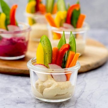 A small glass jar with hummus n the bottom and batons of colourful vegetables standing up in the cup
