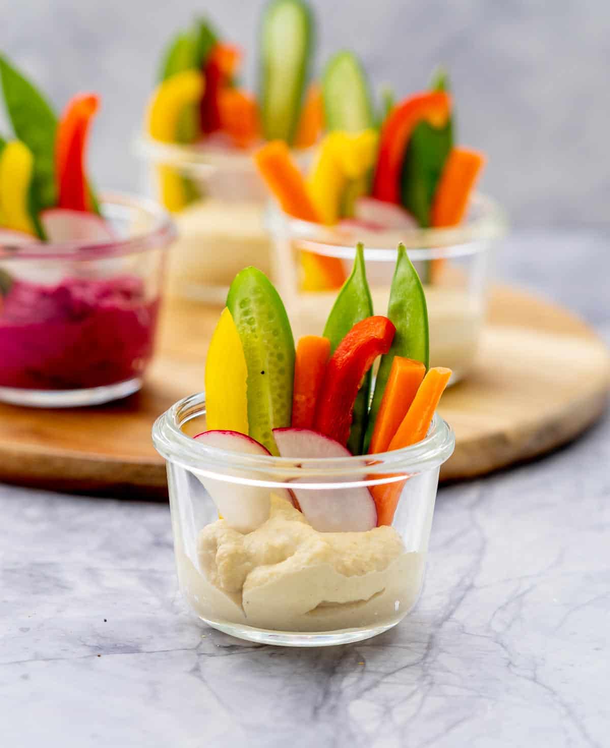 Glass ramekins half filled with hummus and sliced pieces of radishes, carrots, sugar snap peas and cucumber sitting on the bench with a wooden chopping board in the background