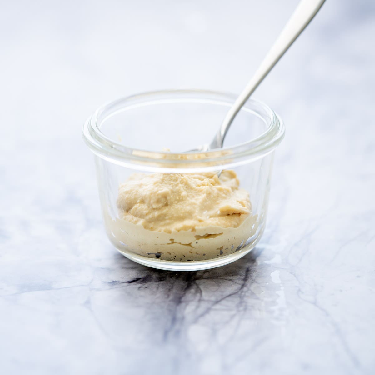 one glass ramekin half filled with hummus with a spoon sitting on the bench with